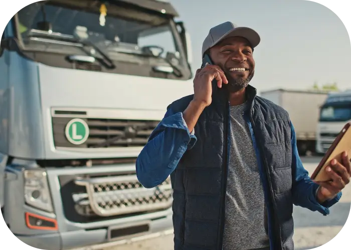 Homem de barba curta usa boné cinza, colete azul marinho acolchoado e camiseta cinza. Ele está em pé, segurando um tablet com a mão esquerda e um celular junto ao ouvido com a direita. Atrás dele há um caminhão prateado com o símbolo verde ‘L’ na frente e outros caminhões estacionados ao fundo. O cenário é um pátio externo iluminado pela luz do dia.