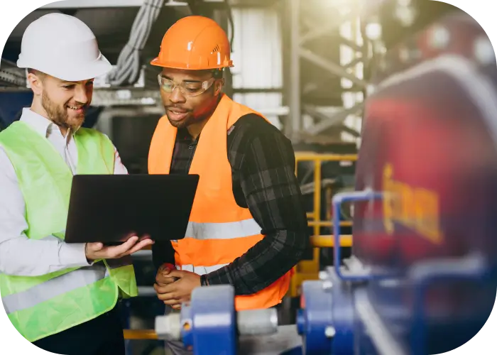 Dois engenheiros em uma área industrial iluminada observam um notebook. Um deles, com capacete branco e colete verde, mostra algo na tela ao colega com capacete e colete laranja. Ambos estão próximos a uma máquina azul com detalhes amarelos e estruturas metálicas ao fundo.