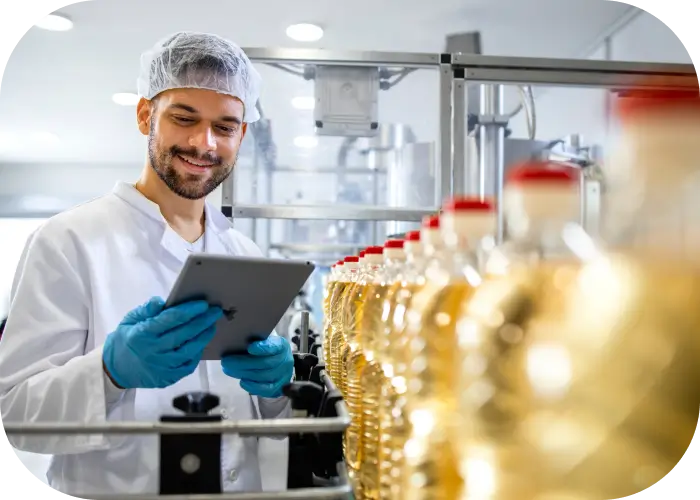 Homem sorrindo com jaleco, touca e luvas azuis analisa informações em um tablet ao lado de uma esteira com garrafas transparentes cheias de líquido dourado, em uma planta industrial limpa e automatizada.