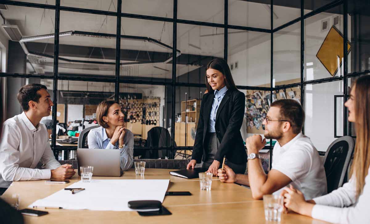 pessoas sentadas em torno de uma mesa de escritório olhando para uma mulher que fala em pé